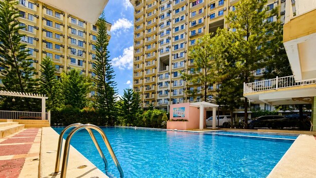 A swimming pool with a metal ladder, surrounded by a building with multiple balconies and trees. - Tranquil 2-Bedroom Apartment with Shared Pool in Davao City (Davao City)