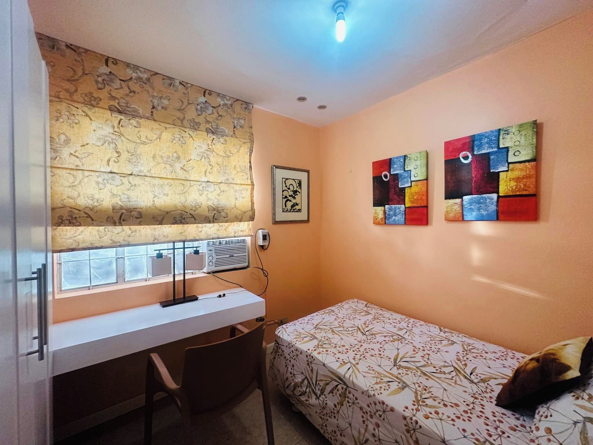 A small room with a desk, a bed, and two paintings on the wall.