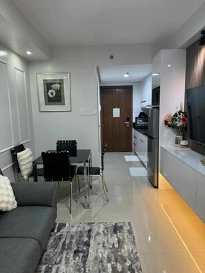 Ein modernes Wohnzimmer mit einer Couch, einem Esstisch mit Stühlen, einer Küchenzeile inklusive Kühlschrank und einem an der Wand befestigten Fernseher.