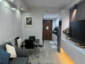 Ein modernes Wohnzimmer mit einer Couch, einem Esstisch mit Stühlen, einem Flachbildfernseher und einer Küchenzeile inklusive Kühlschrank und Mikrowelle.