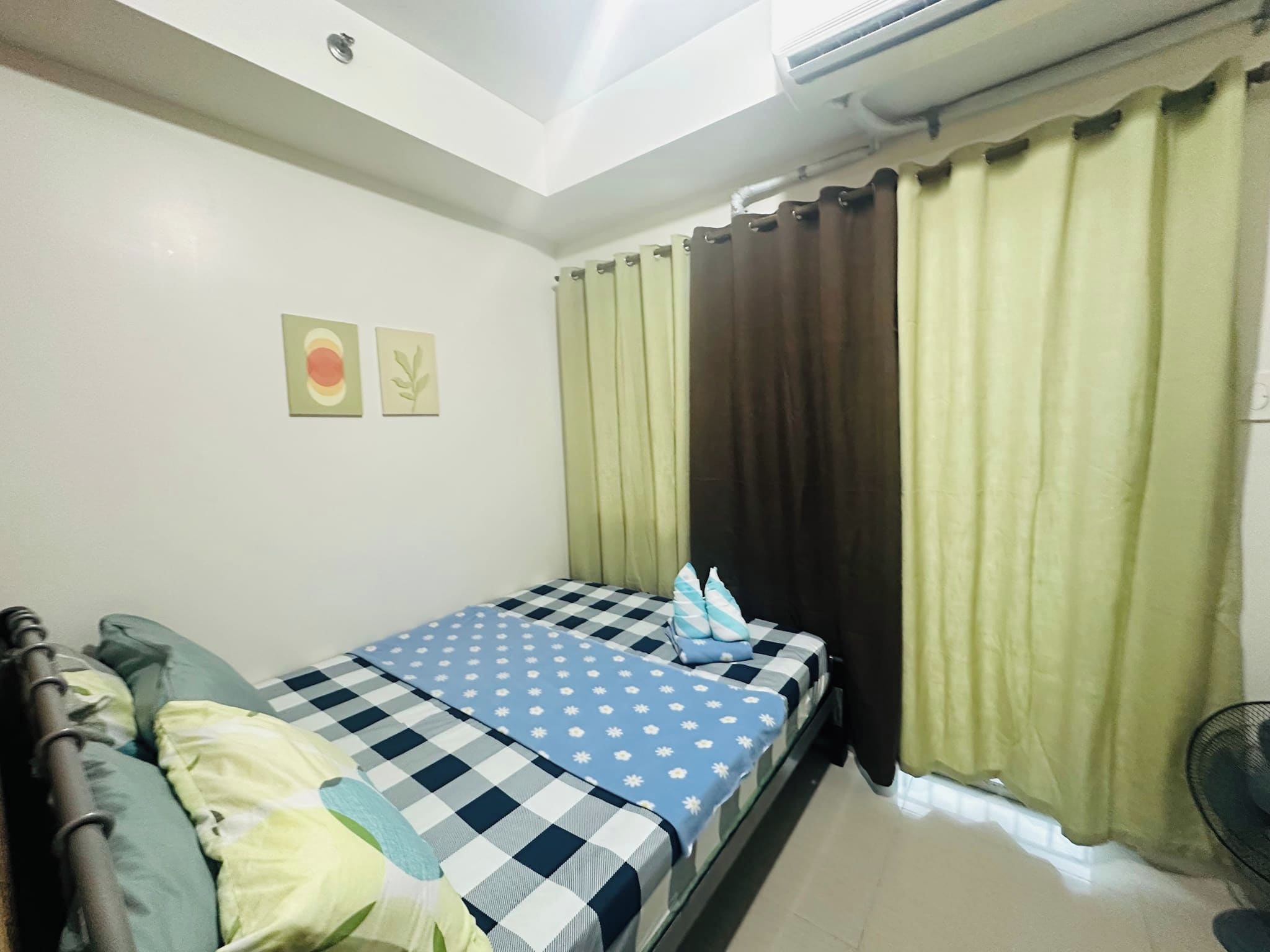 A single bed with a checkered bedspread, a pillow, and a blanket. There is a window with curtains, a wall-mounted air conditioning unit, and two wall art pieces.