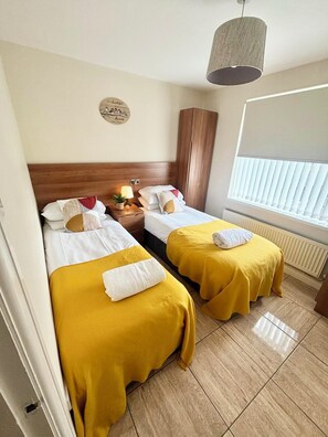 A hotel room with two beds, a wooden headboard, a bedside table with a lamp, a window with blinds, and a wall decoration. - Beautiful 3 bed, 2 bath apartment with free on-road parking, WiFi & bills inc. (Parade)