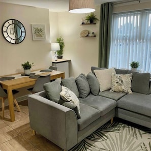 A modern living room with a grey sectional sofa, a dining table with chairs, a round wall clock, and a window with blinds. - Beautiful 3 bed, 2 bath apartment with free on-road parking, WiFi & bills inc. (Parade)