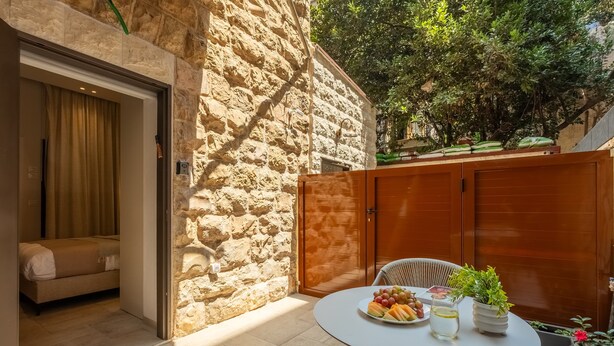 Una habitación de paredes de piedra con una cama, una mesa con frutas y un vaso de agua, y vistas a la vegetación exterior.
