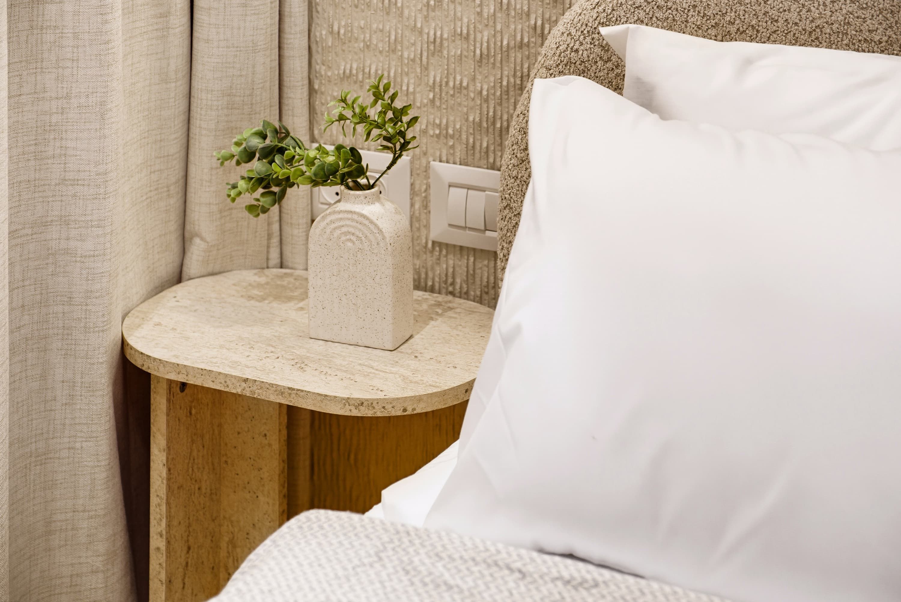 A bed with white bedding, a round wooden nightstand with a white vase containing green plants, and a white switch on the wall.