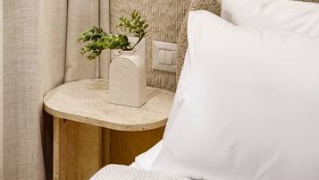 A bed with white bedding, a round wooden nightstand with a white vase containing green plants, and a white switch on the wall.
