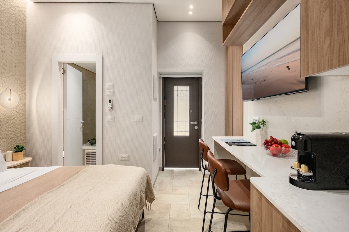 A modern hotel room with a bed, a desk with a computer, a coffee maker, and a TV.