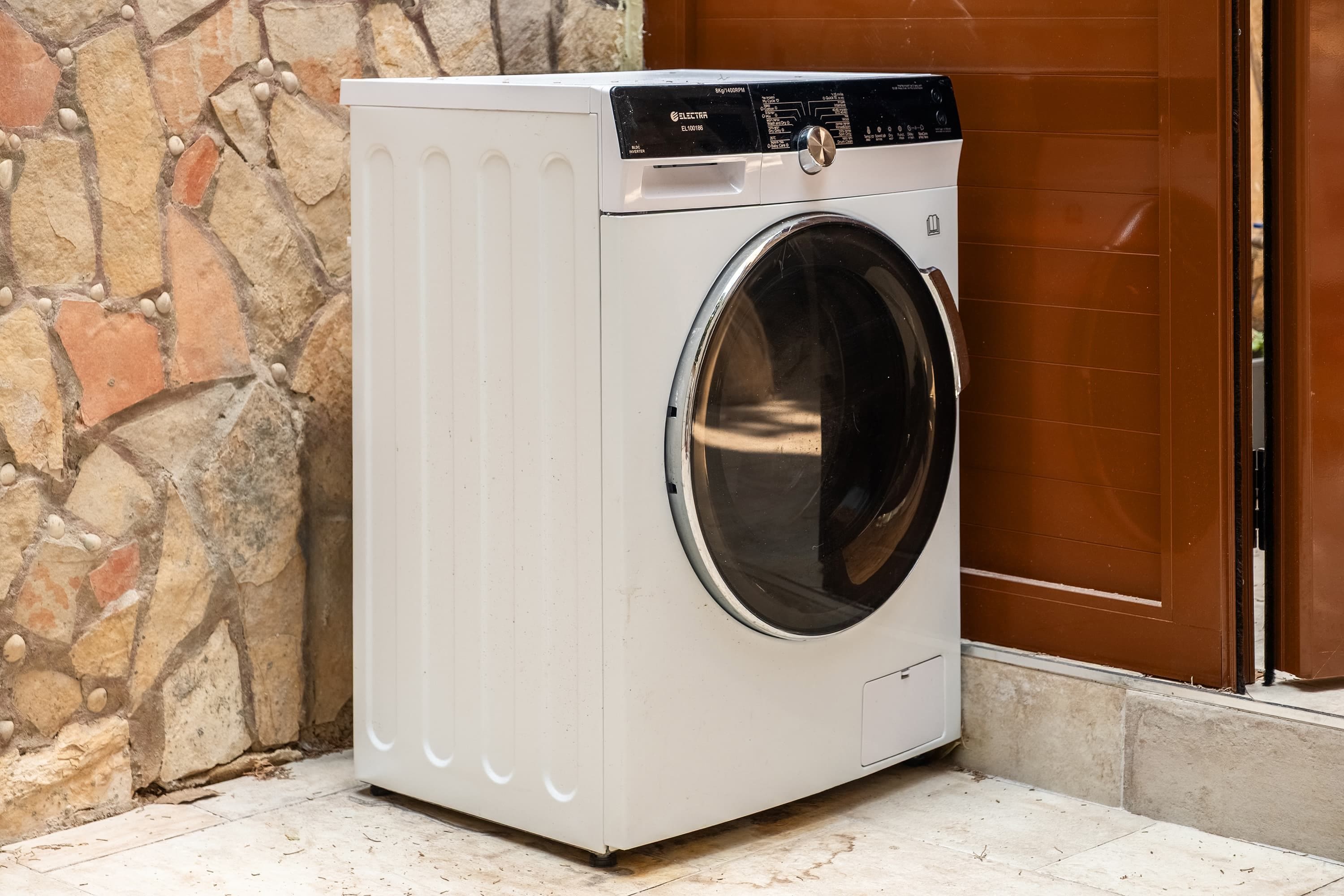 A front-loading washing machine with a control panel, placed against a stone wall and a brown door.