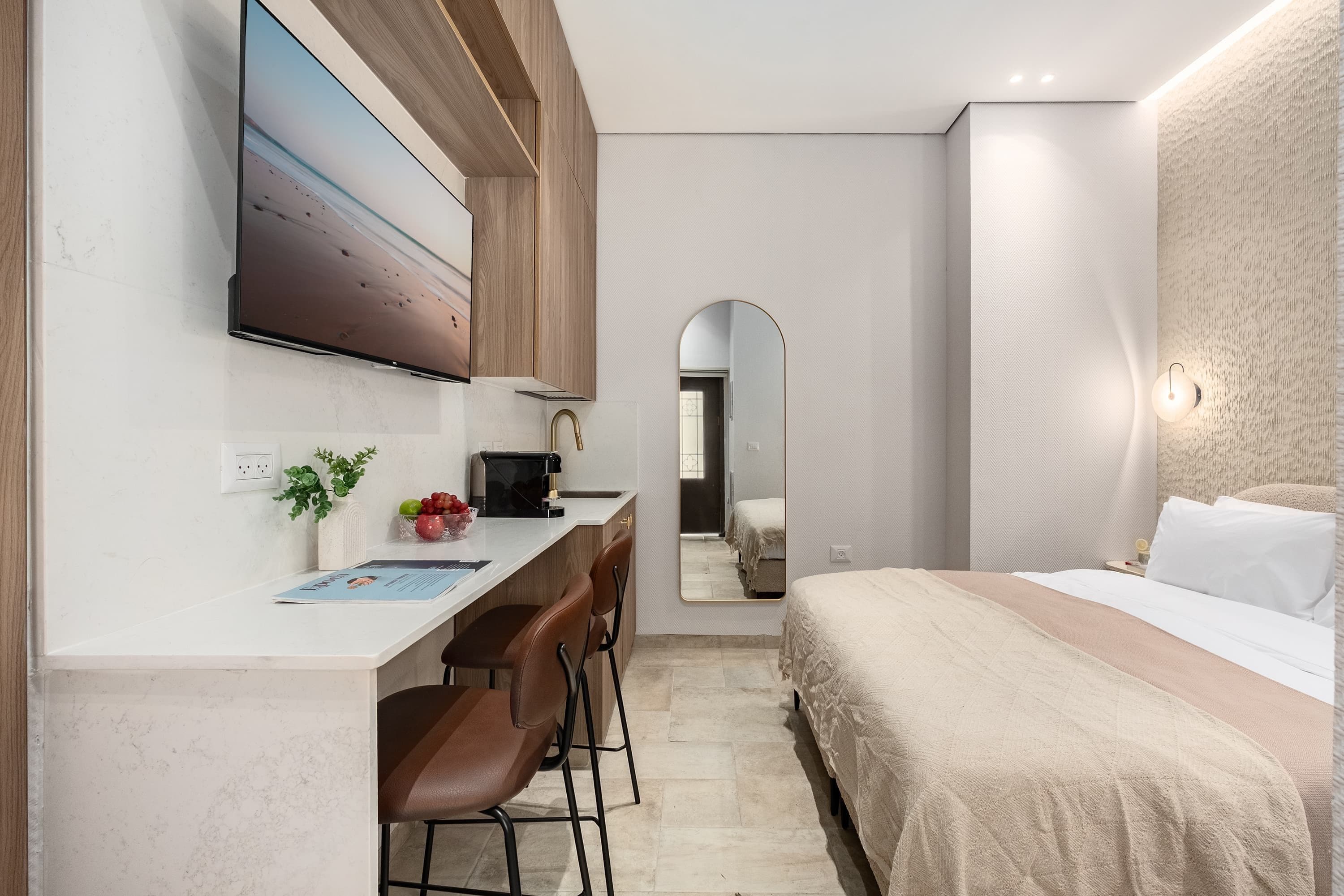 A compact hotel room with a bed, a desk with a chair, a TV mounted on the wall, and a mirror.