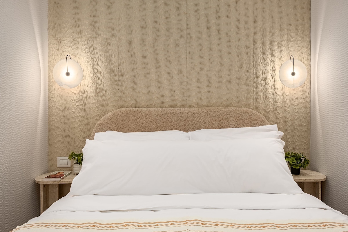 A neatly made bed with white linens and a patterned throw, a bedside table with a book and a plant, and a textured wall with two wall-mounted lights.