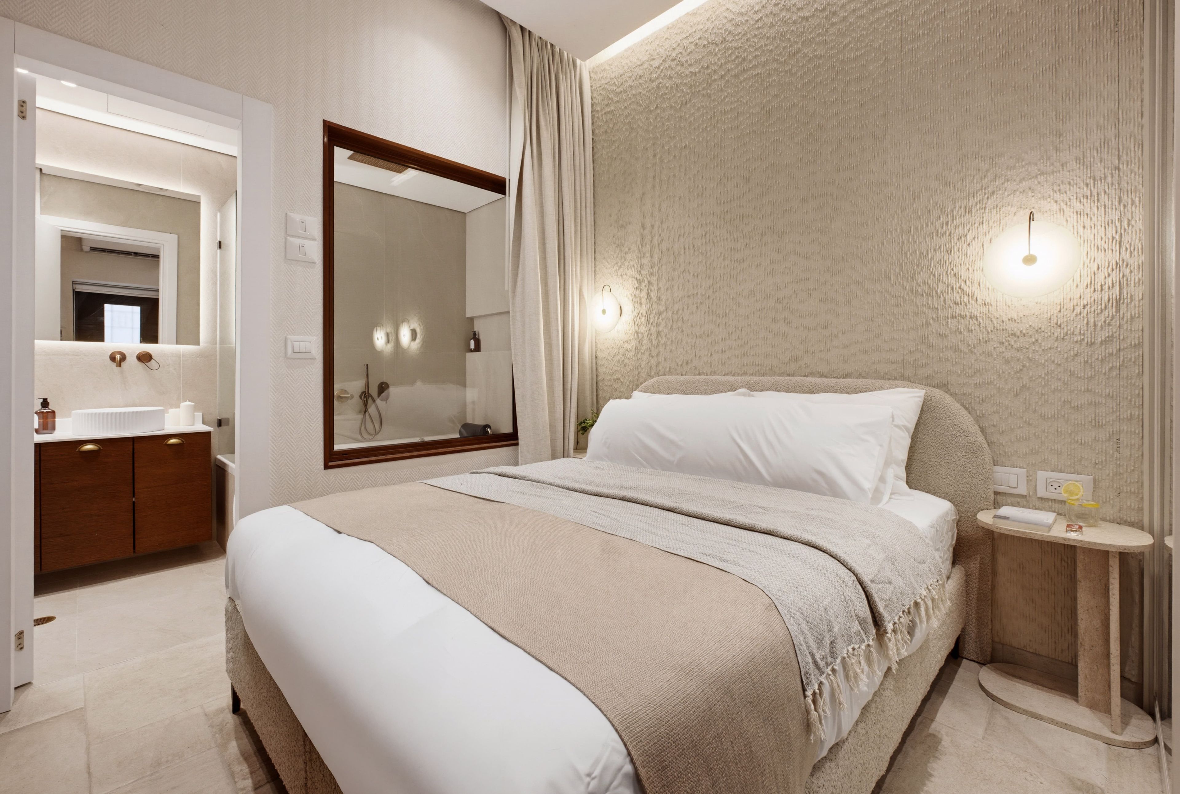 Habitación de hotel con una cama grande, un baño con espejo y una mesita de noche con lámpara.