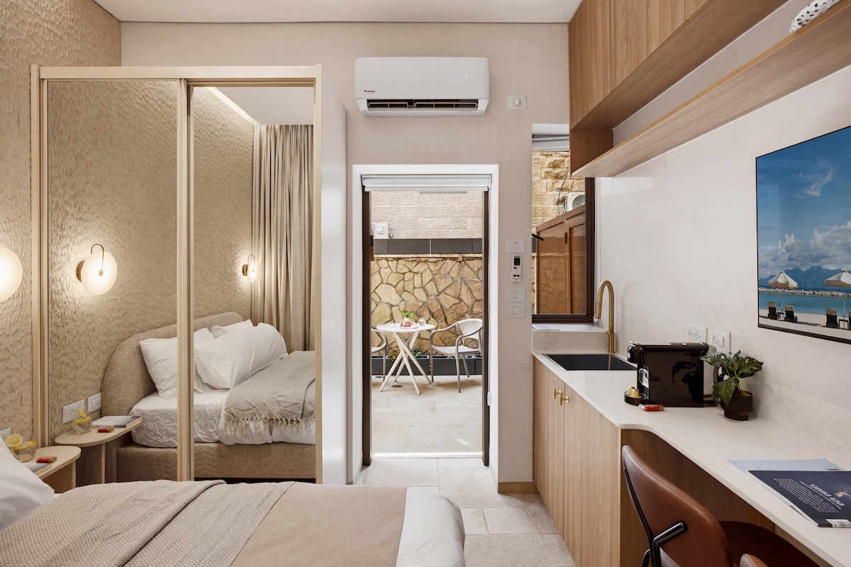 A modern hotel room with a bed, bedside table, and a view of a dining area.