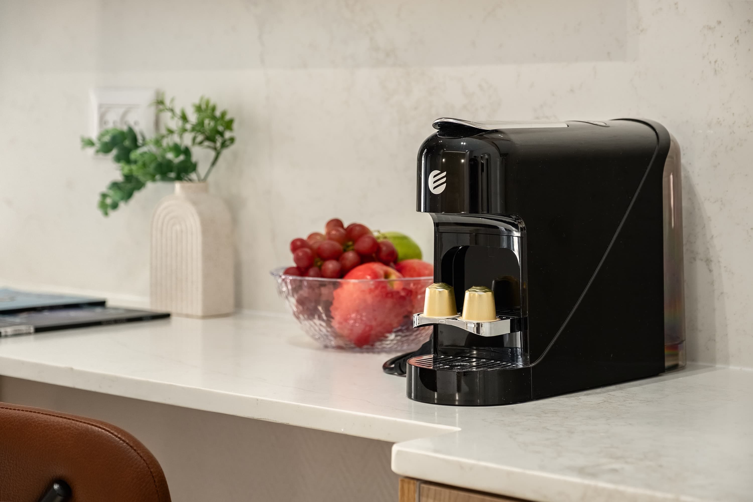 A kitchen with a coffee maker, a bowl of fruit, and a vase with greenery.