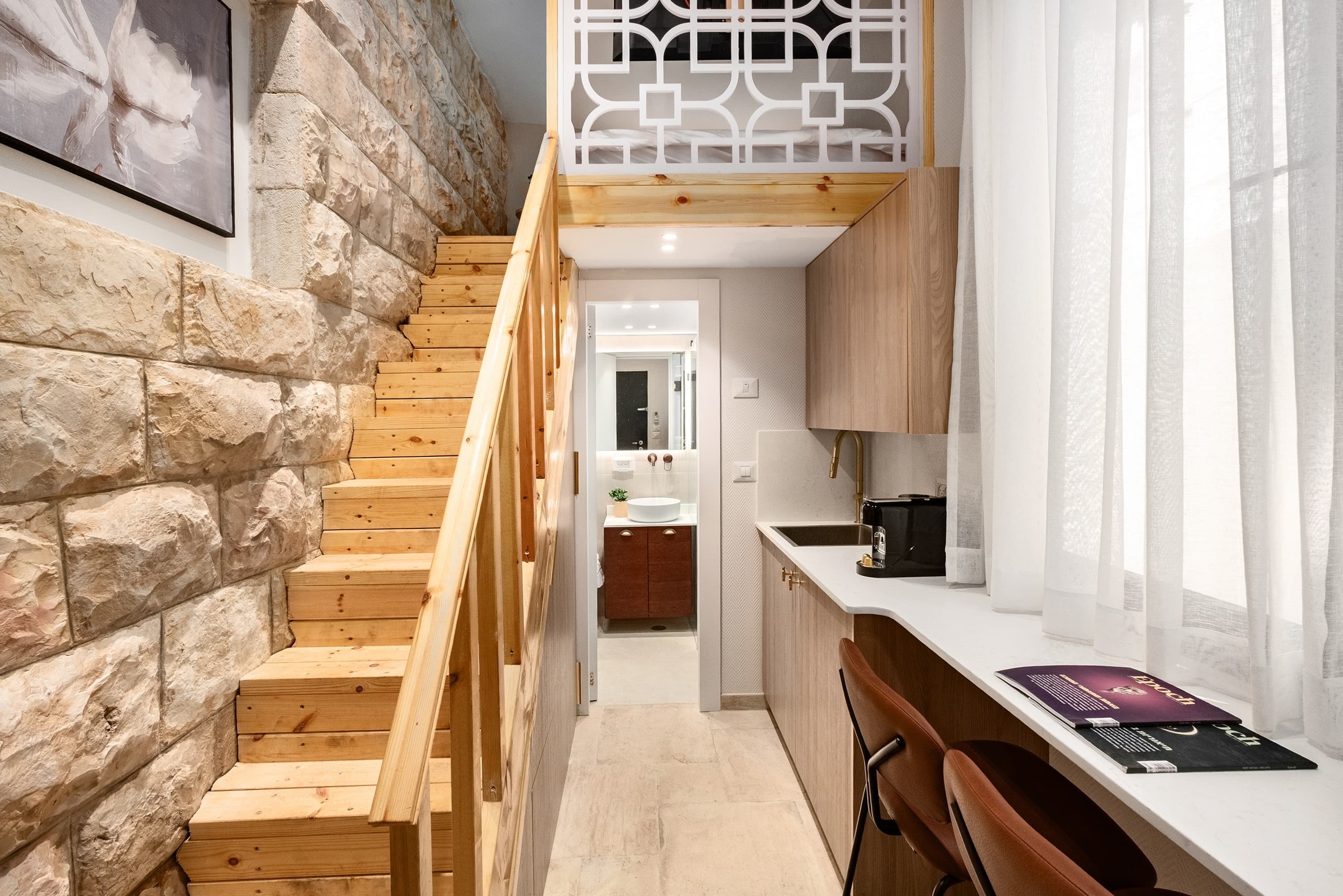 Una escalera conduce a un dormitorio tipo loft con piso de madera, pared de piedra y un panel geométrico blanco.