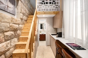 Una escalera conduce a un dormitorio tipo loft con piso de madera, pared de piedra y un panel geométrico blanco.