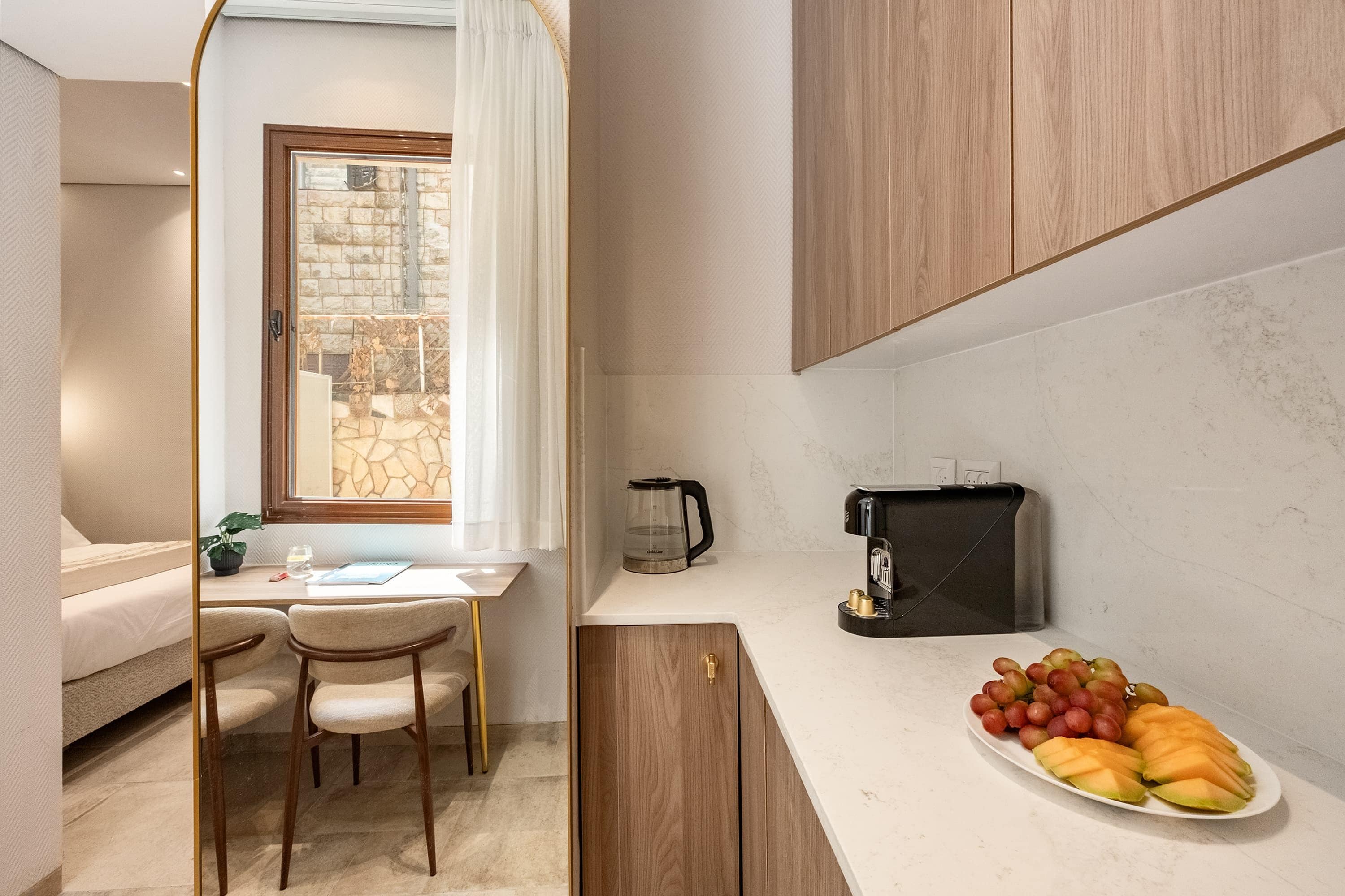 A compact kitchen with a coffee maker, a fruit platter, and a view of a stone wall through a window.