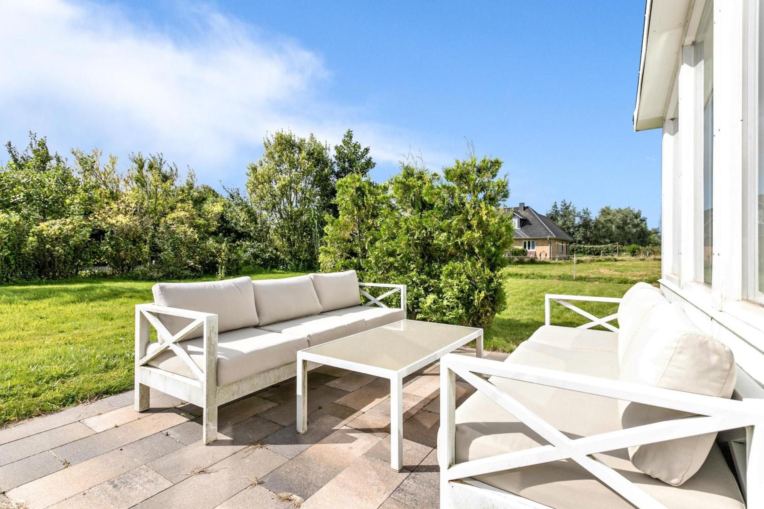 Ein Sitzbereich im Freien mit einer Couch und Stühlen, einem Tisch und Blick auf ein Haus und Grünpflanzen im Hintergrund.