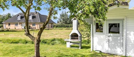 Un entorno rústico al aire libre con un horno de piedra tradicional, una estructura de madera blanca y una casa al fondo.