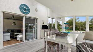 A modern living room with a white brick wall, a large anchor-themed clock, a sliding glass door leading to a balcony, and a dining area with a white table and chairs. - "Ebbe" Holiday Apartment with Private Terrace, Shared Garden, and Wi-Fi (Rodenäs)