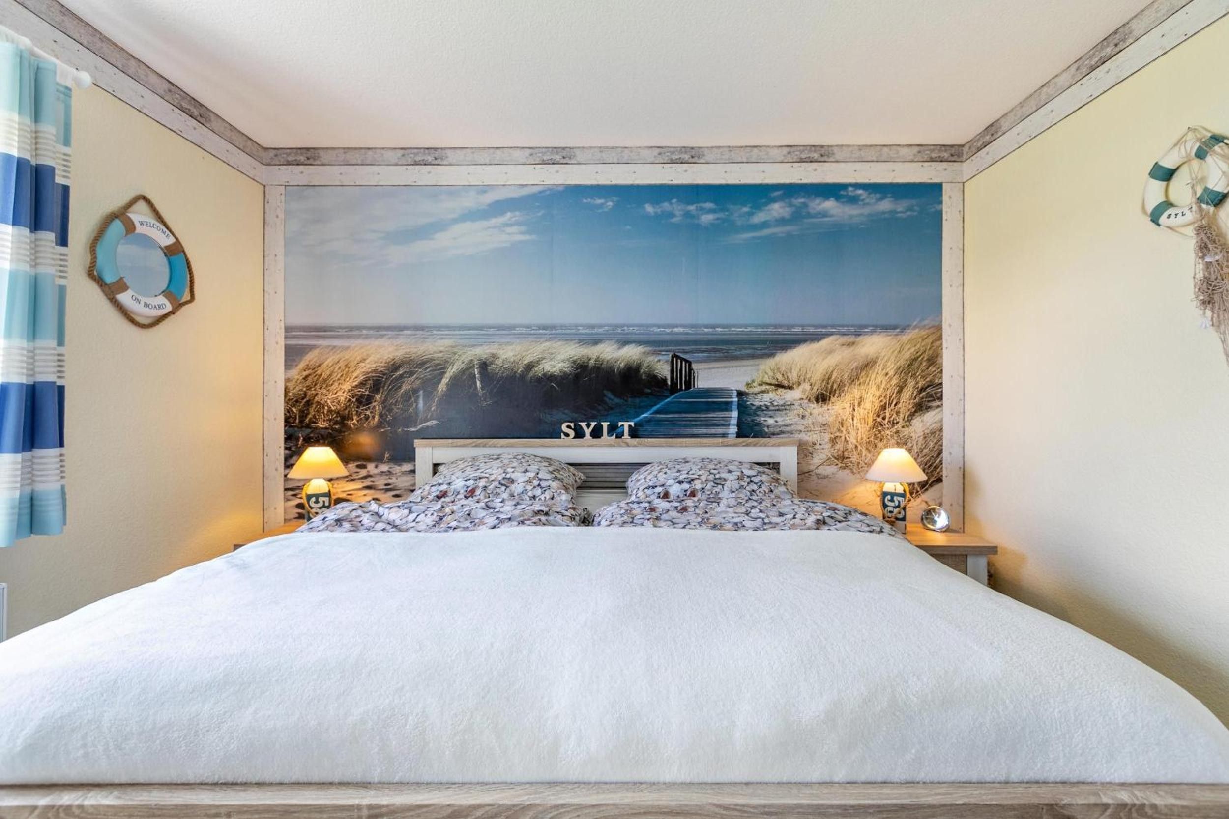 Een slaapkamer met een grote muurschildering in strandthema, een bed met witte lakens, twee kussens en een nachtkastje met lampen.