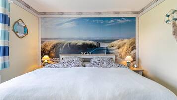 Een slaapkamer met een grote muurschildering in strandthema, een bed met witte lakens, twee kussens en een nachtkastje met lampen.