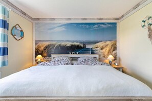 Ein Schlafzimmer mit einer großen Wandmalerei im Strandthema, einem Bett mit weißer Bettwäsche, zwei Kissen und einem Nachttisch mit Lampen.