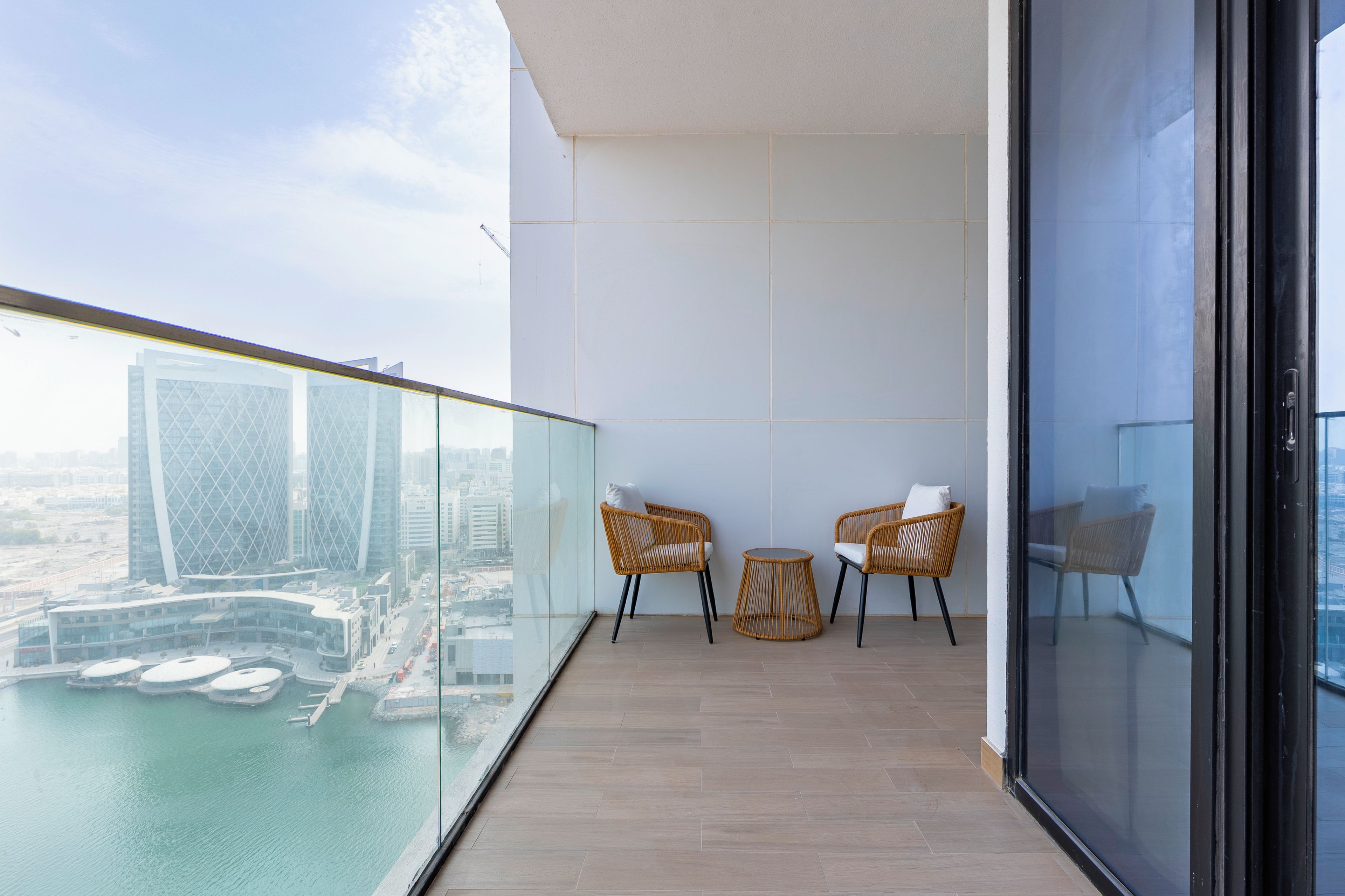 A balcony with a glass railing, two wicker chairs, a small round table, and a view of a cityscape with buildings and a body of water.