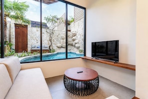 A modern living room with a large window, a pool, a sofa, a round table, and a flat-screen TV. - NEW 1BR Villa Svargi 1 Surf and Sanctuary Private pool (Pecatu, South Kuta, Badung Regency, Bali 80361)