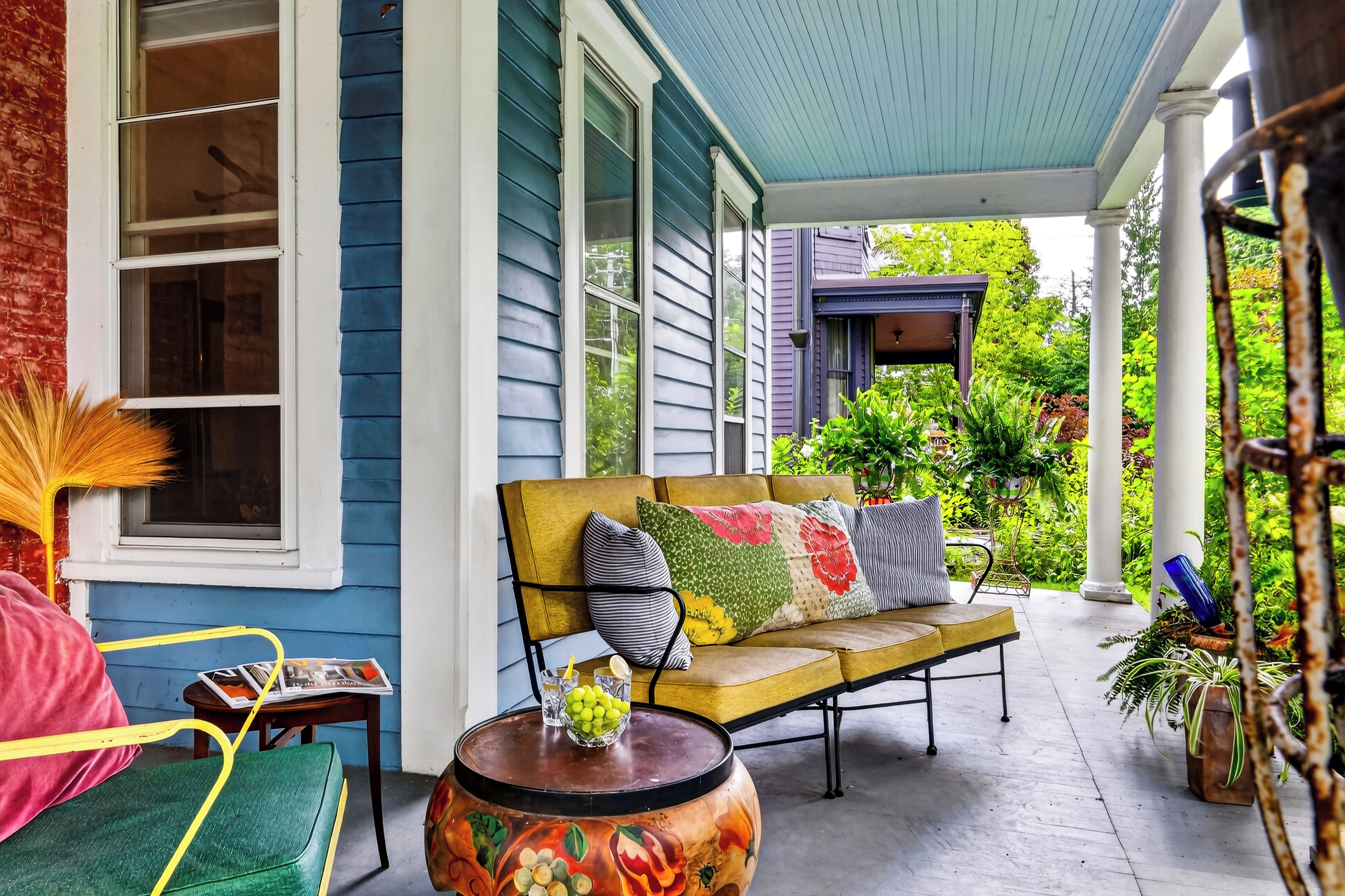 A porch with a yellow sofa, a round table with fruit, and potted plants.