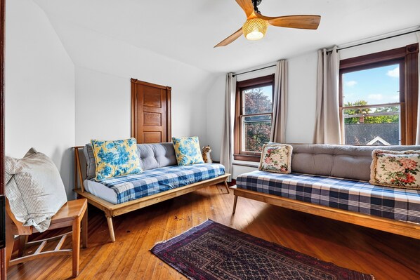 Une pièce avec un plancher en bois, deux canapés ornés de coussins à motifs, une chaise en bois, un ventilateur de plafond et une fenêtre avec des rideaux.