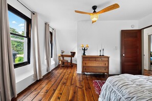 Une chambre avec un plancher en bois, un lit, une commode, un ventilateur de plafond et une fenêtre avec des rideaux.