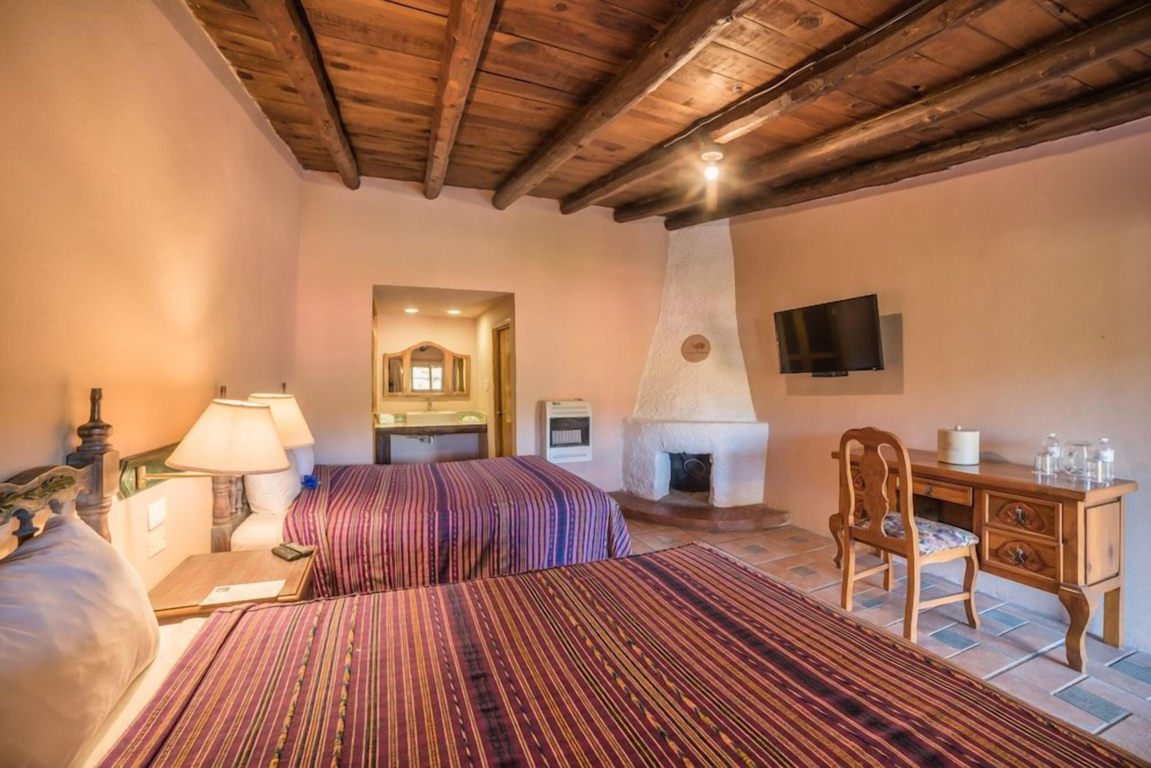 A bedroom with a wooden ceiling, a bed with striped bedding, a desk with a chair, a television, and a fireplace.