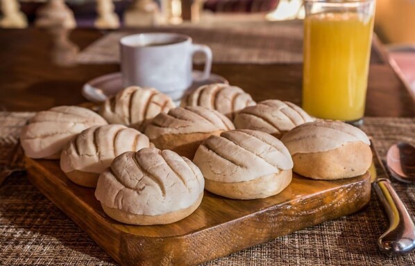 A wooden board with several round, textured pastries, a white cup, and a glass of orange juice on a table. - Hotel Barrancas del Cobre (Urique)
