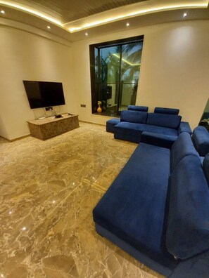 A modern living room with a flat-screen TV, a marble floor, and a large window with a view of palm trees. - 23 West, Malavli (Pune)
