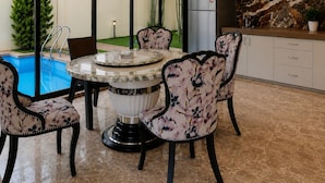 A dining area with a round table, chairs, a ceiling light fixture, and a view of a swimming pool and greenery outside. - 23 West, Malavli (Pune)