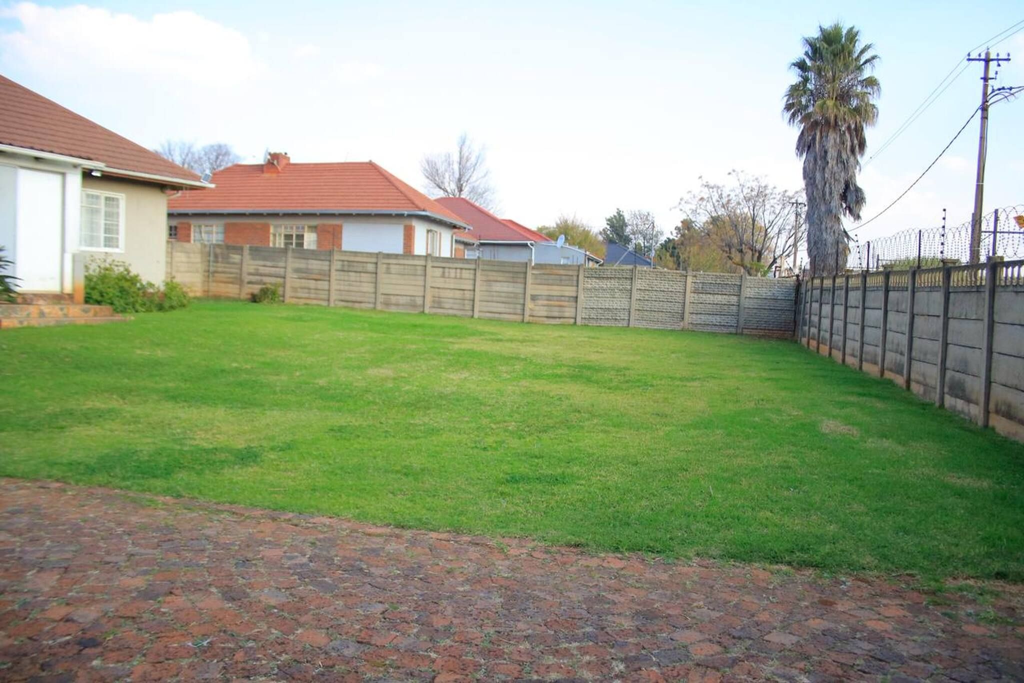 Un patio trasero residencial con camino de entrada pavimentado, cerca de madera y una palmera.