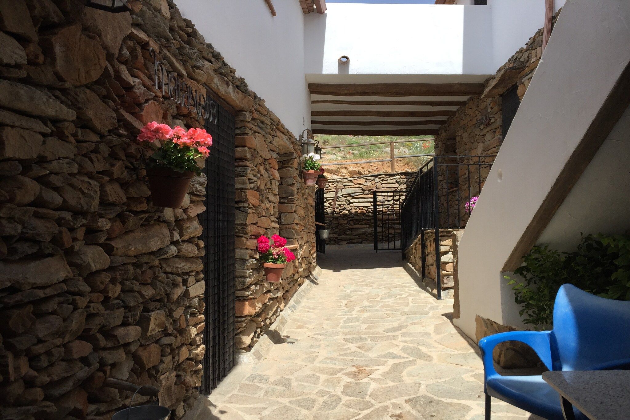 Un estrecho sendero de piedra que conduce a un espacio al aire libre cubierto, con una silla azul y plantas en macetas.