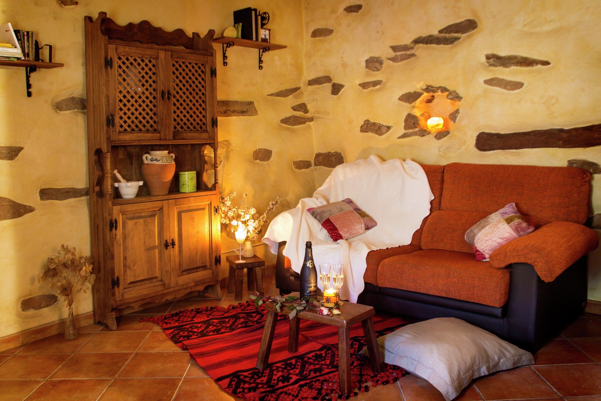 Uma sala de estar acolhedora com parede de pedra, um armário de madeira com cerâmica, um tapete vermelho estampado e um sofá laranja confortável.