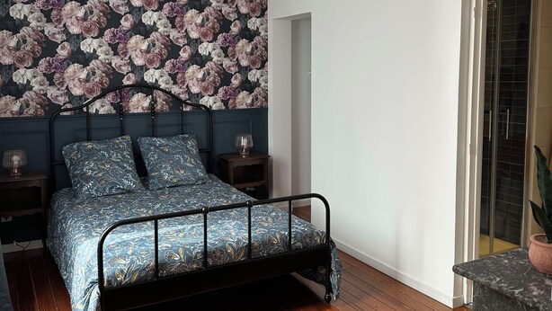 Un dormitorio con cama, mesitas de noche, papel tapiz floral, suelo de madera y vistas por una puerta.