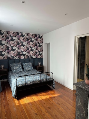 Un dormitorio con cama, mesitas de noche, papel tapiz floral, suelo de madera y vistas por una puerta.