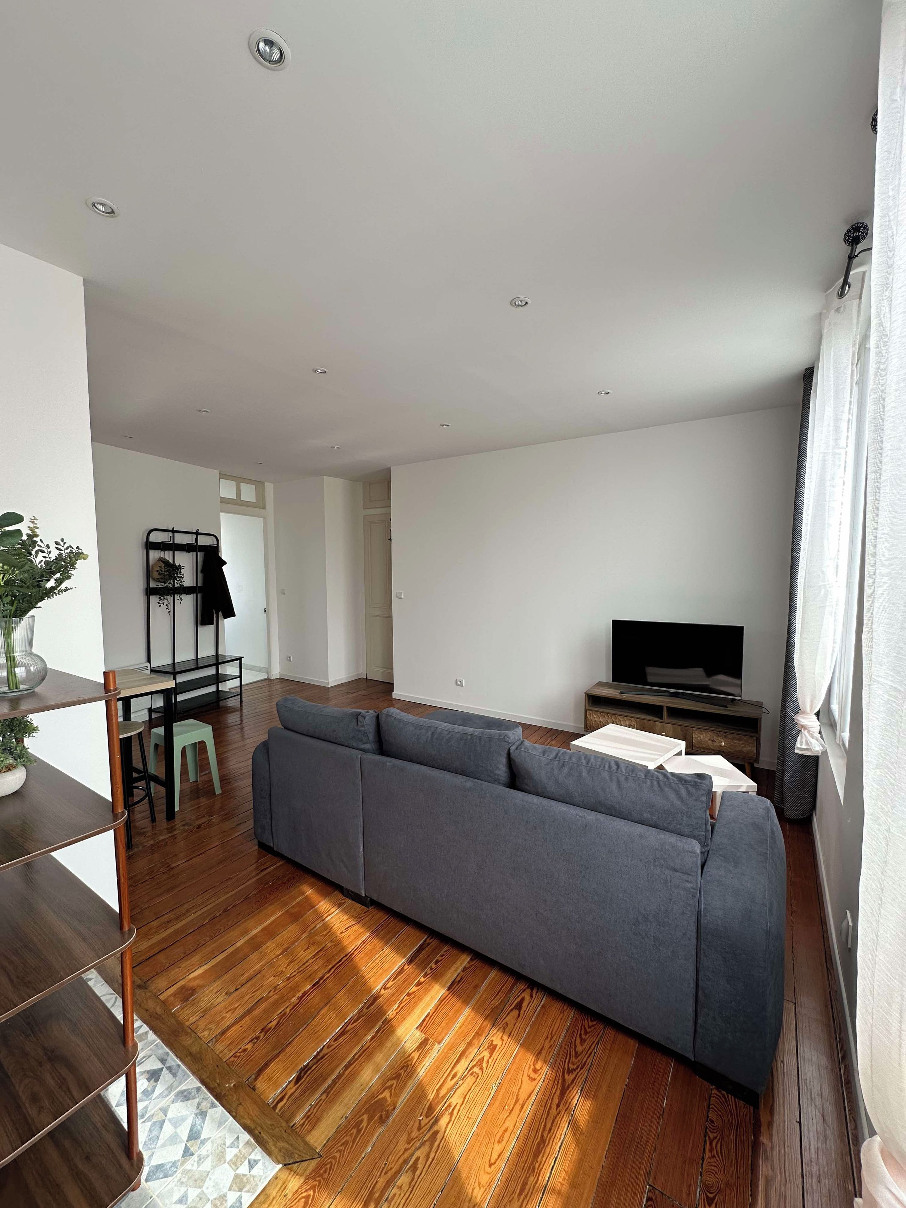 Een moderne woonkamer met een grijze bank, houten vloeren, een televisie en een eethoek met een kleine tafel en stoelen.