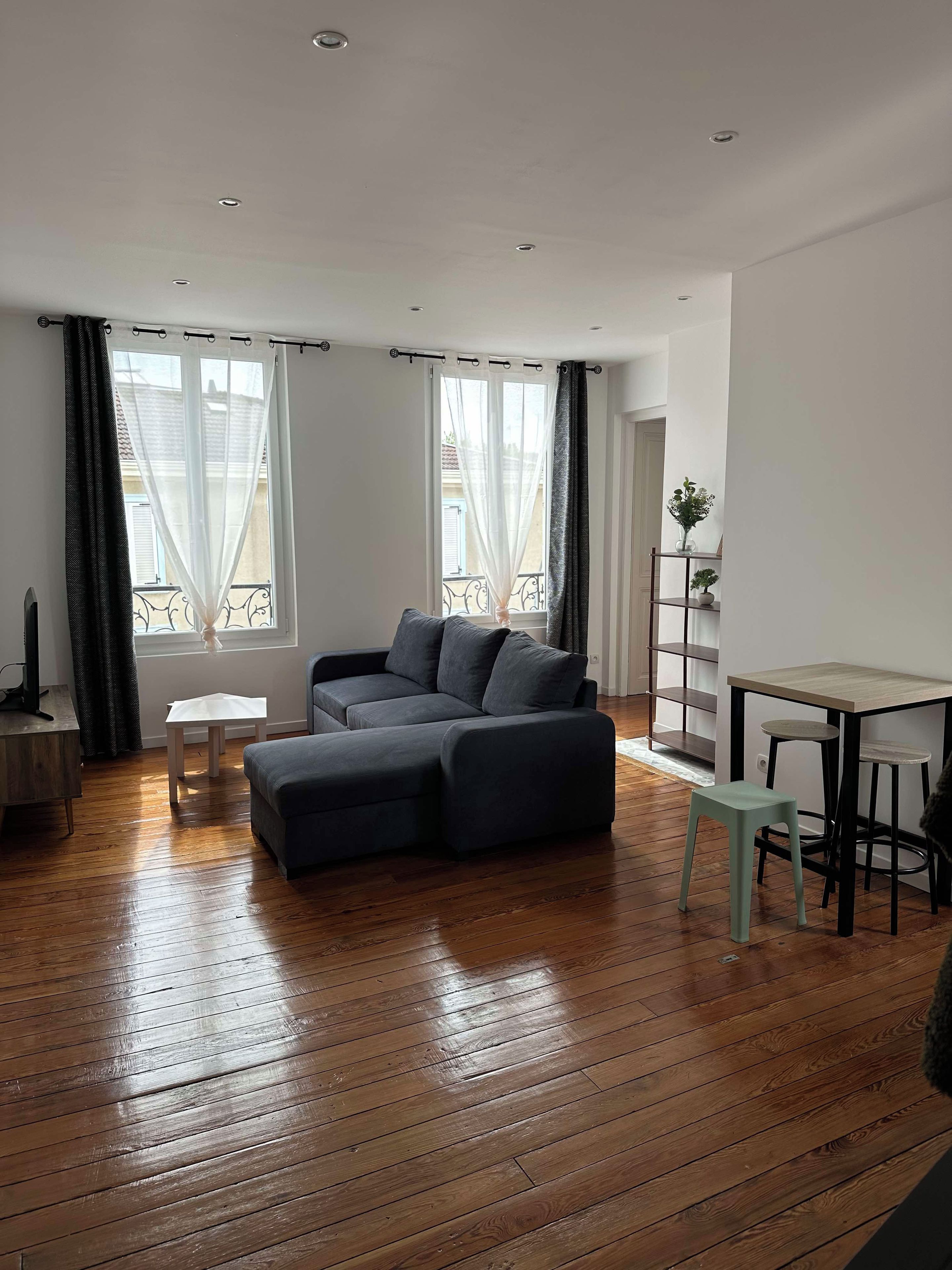 A modern living room with a dark grey sectional sofa, a small white table, and a dining area with a light green stool.