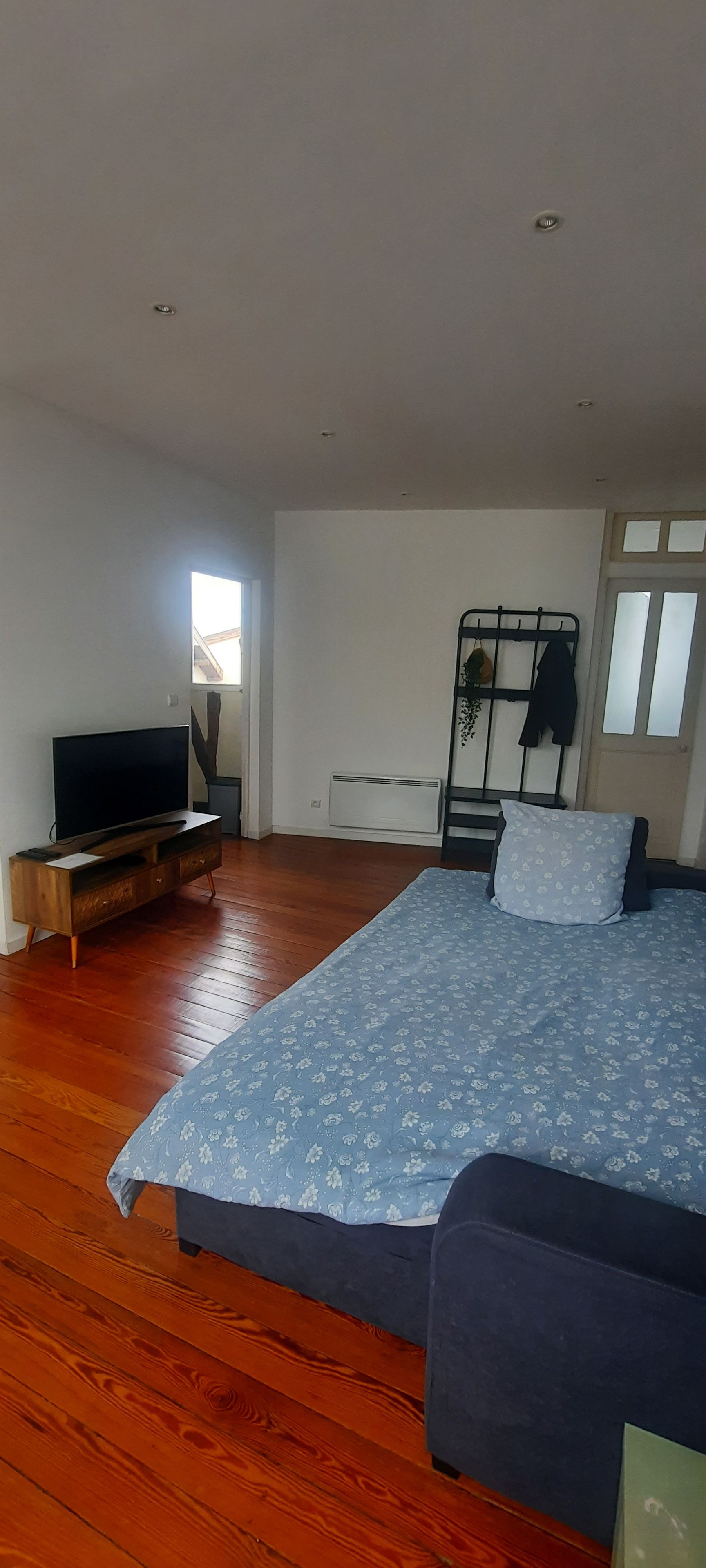 Een slaapkamer met een bed, een televisie op een houten standaard, een radiator en een raam met zonwering.