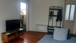 Un dormitorio con una cama, un televisor sobre un soporte de madera, un radiador y una ventana con persianas.