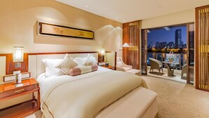 Habitación de hotel con una cama grande, una mesita de noche, un escritorio, una silla y un balcón con vistas a la ciudad.