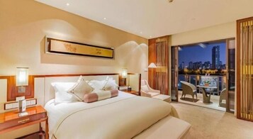 Habitación de hotel con una cama grande, mesitas de noche, vistas de la ciudad y un balcón con asientos.