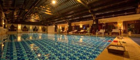An indoor swimming pool with a checkered tile floor, lounge chairs, and a wooden deck area.