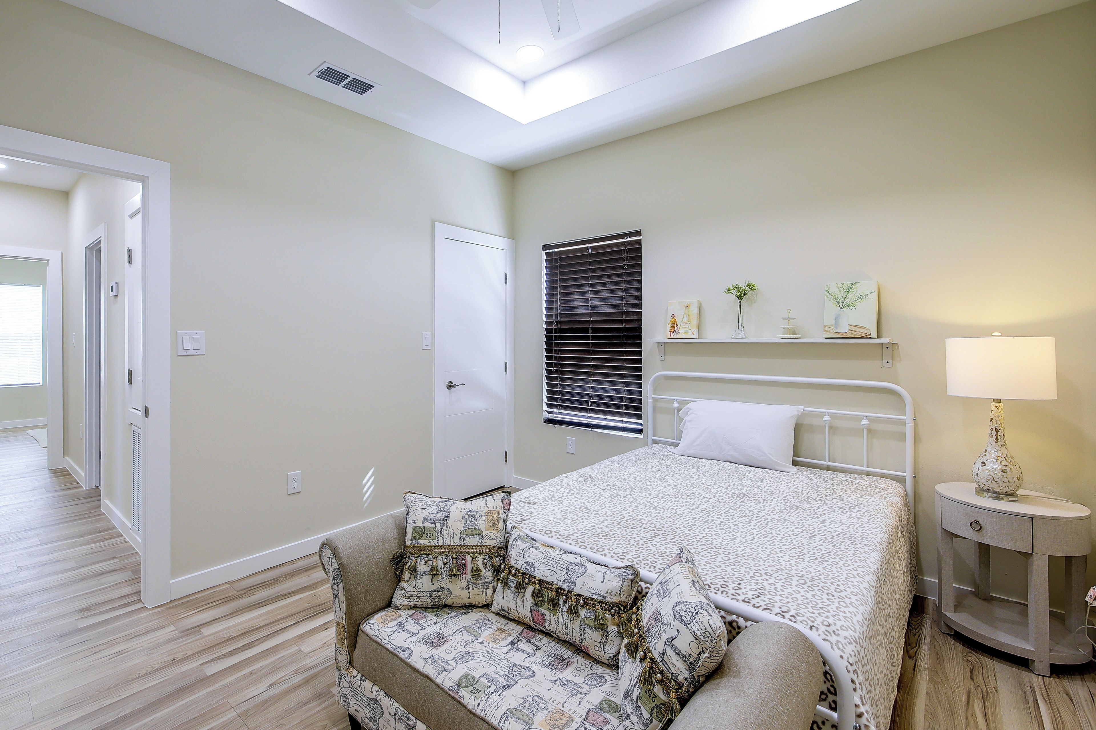 A bedroom with a bed, a nightstand, a lamp, a window with blinds, a door, and a hallway.