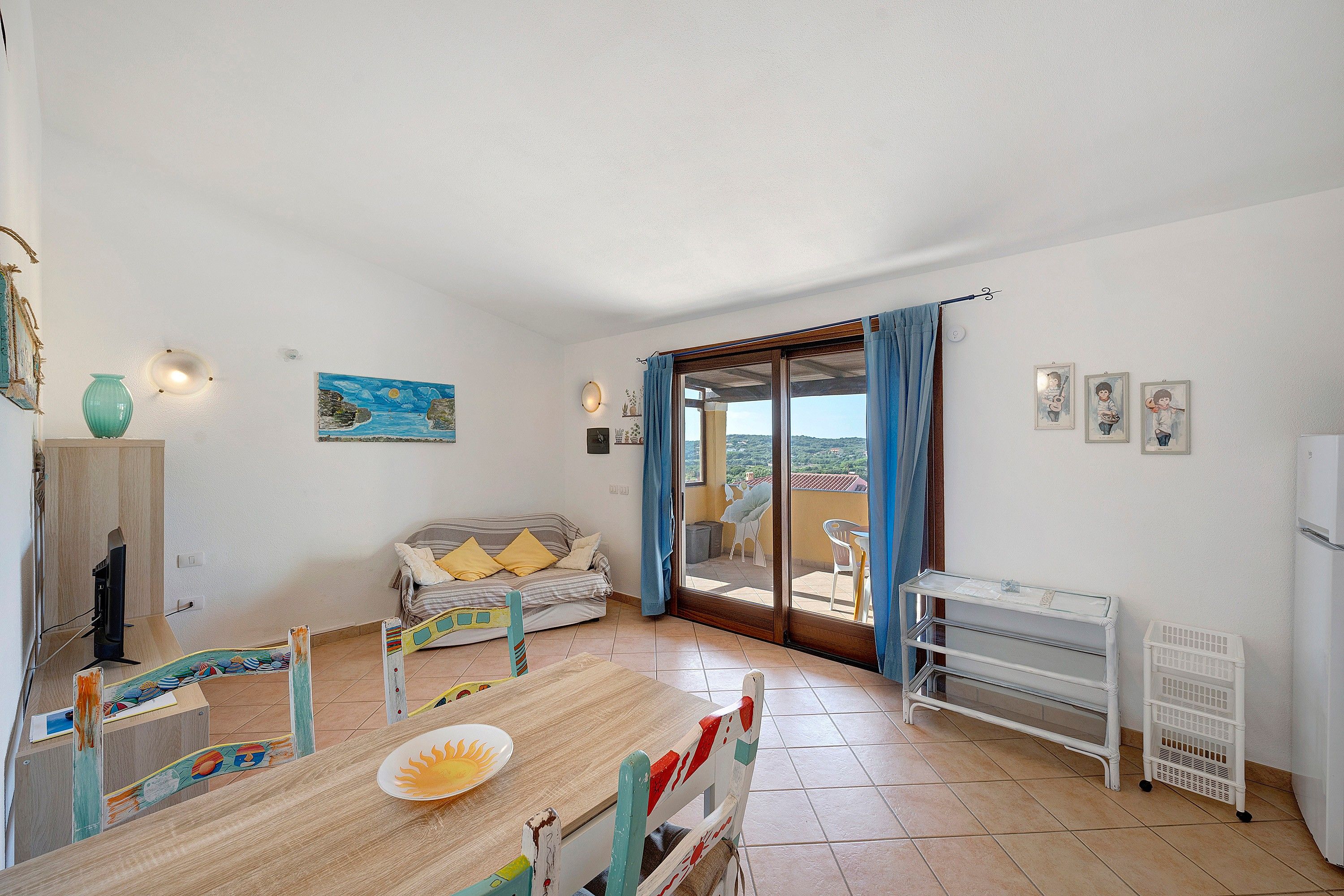 Quarto com mesa de jantar, sofá, televisão e vista através de uma porta de vidro.