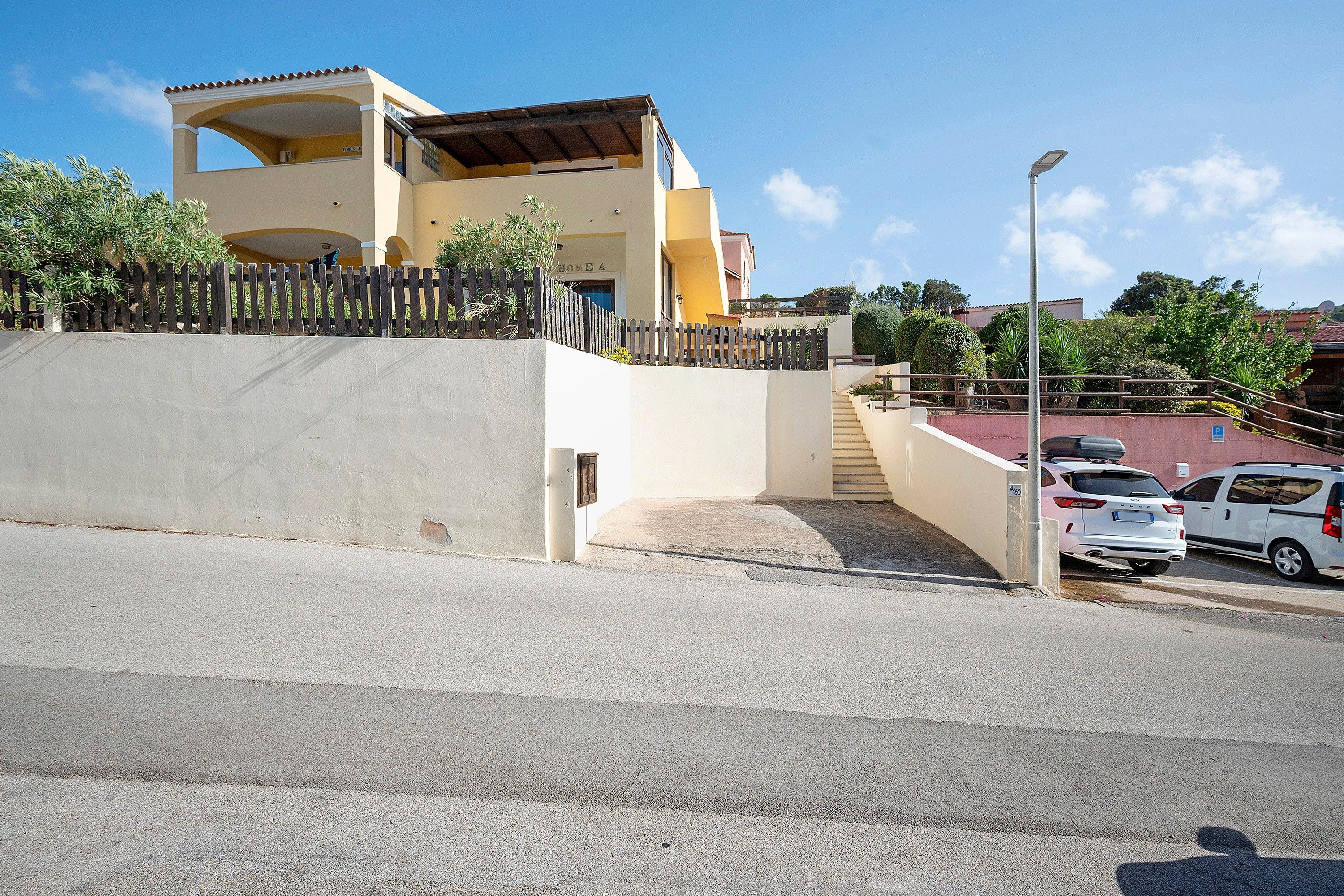 A two-story building with a flat roof, a wooden fence, and a paved driveway leading to a parking area with cars.